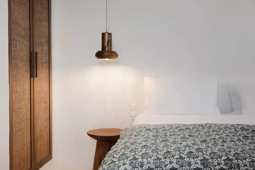 Bett mit gemusterter Tagesdecke neben einem kleinen Holztisch und einer hängenden Lampe.
