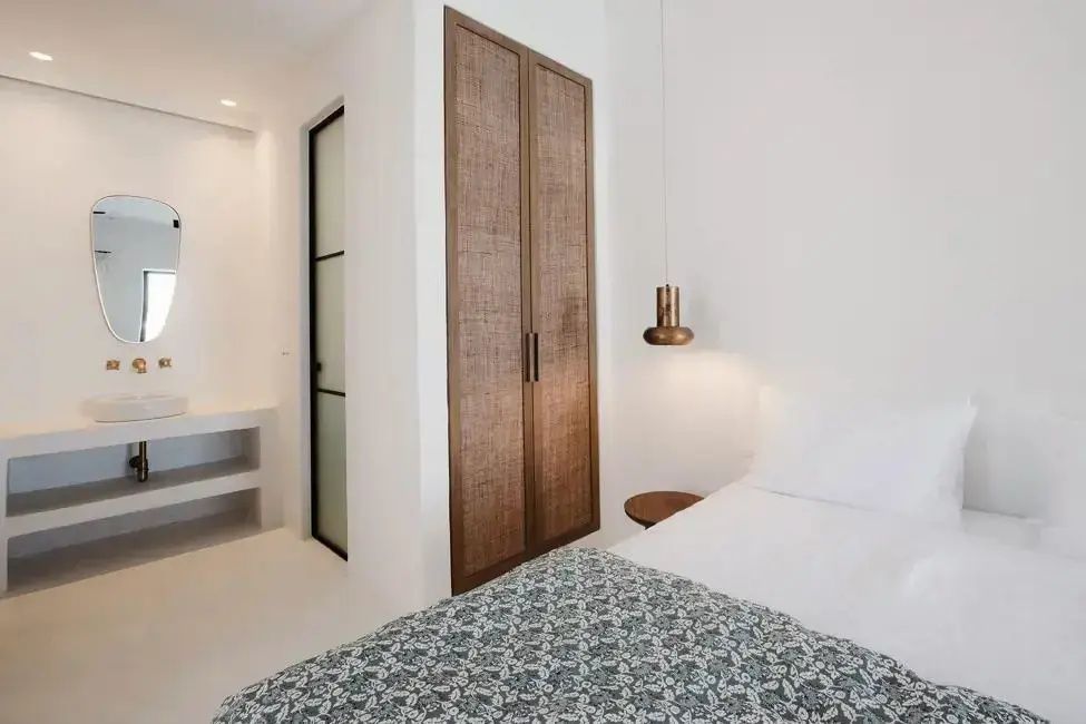 Hotelzimmer mit weißem Bett, einem Holzschrank mit Geflecht und moderner Hängelampe.