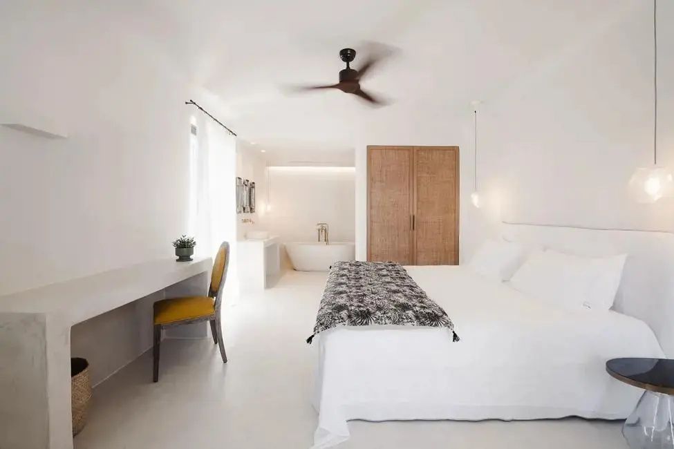 Helles Hotelzimmer mit weißem Doppelbett, Holzschrank, Deckenventilator und offener Badewanne.