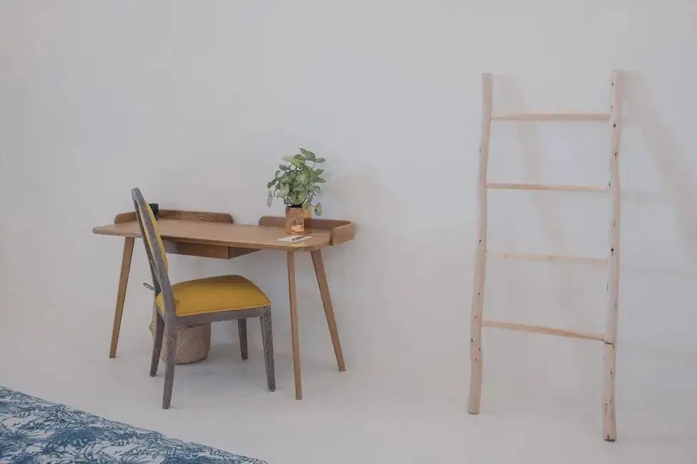 Holzschreibtisch mit einem gelben Stuhl und einer dekorativen Holzleiter an einer weißen Wand.
