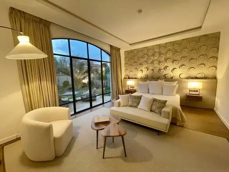 Hotelzimmer mit Doppelbett, beigem Sofa und großer Rundbogenfensterfront mit Blick in den Garten.