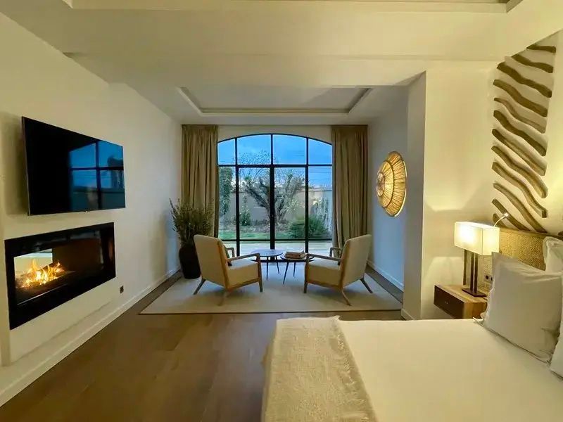 Helles Hotelzimmer mit Kamin, Fernseher, zwei Sesseln und Blick in den Garten.