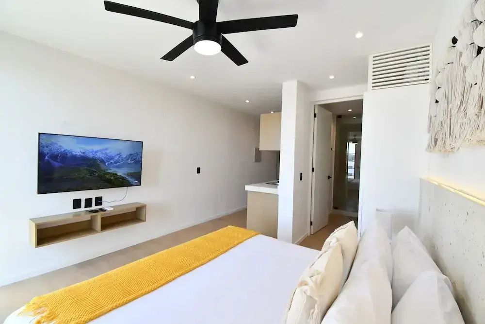 Helles Schlafzimmer mit Doppelbett, gelber Decke, Deckenventilator und Fernseher an der Wand.