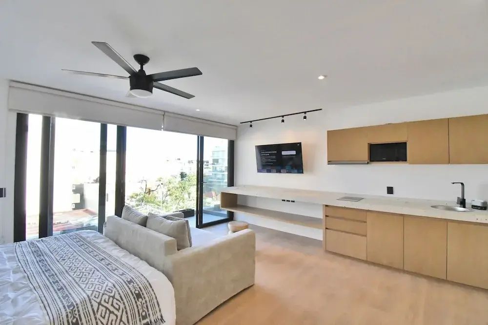 Modernes Zimmer mit Küchenzeile, Wand Fernseher, hellen Holzmöbeln und großer Glasfront zum Balkon.