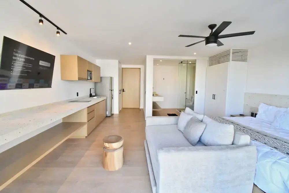 Helles Studio mit Küchenzeile, Kühlschrank, hellen Holzelementen, Sofa und gläserner Badezimmerwand.