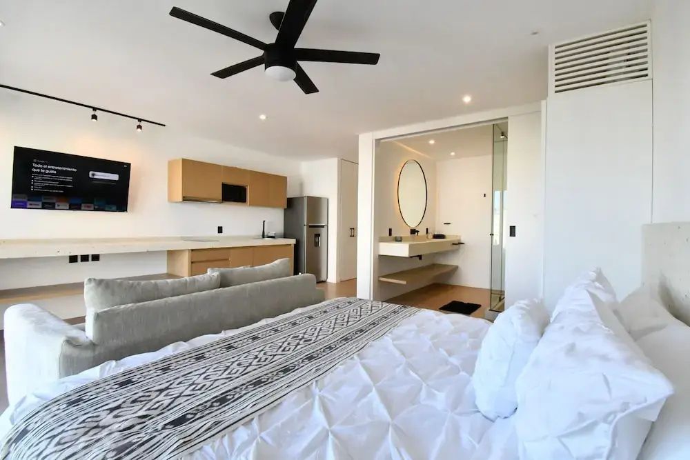 Studiozimmer mit Doppelbett, hellem Sofa, Deckenventilator und angrenzender Küchenzeile.
