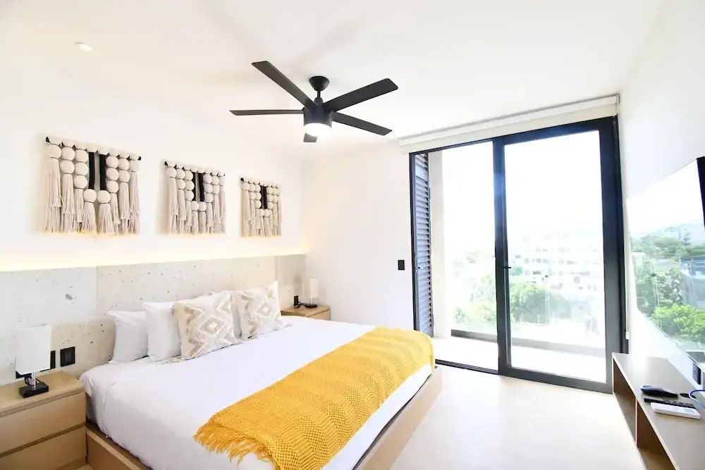 Doppelbett mit gelber Tagesdecke, Makramee-Wanddeko und bodentiefem Fenster zum Balkon.