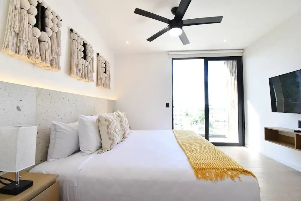 Bett mit weißen Kissen, gelber Decke und Wanddekoration neben einer großen Glasfront.