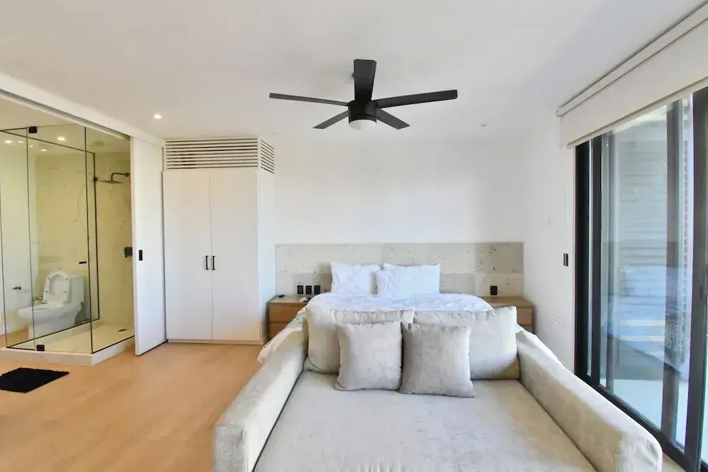 Doppelbett mit Kissen, helles Sofaelement davor, weißer Kleiderschrank und Deckenventilator.