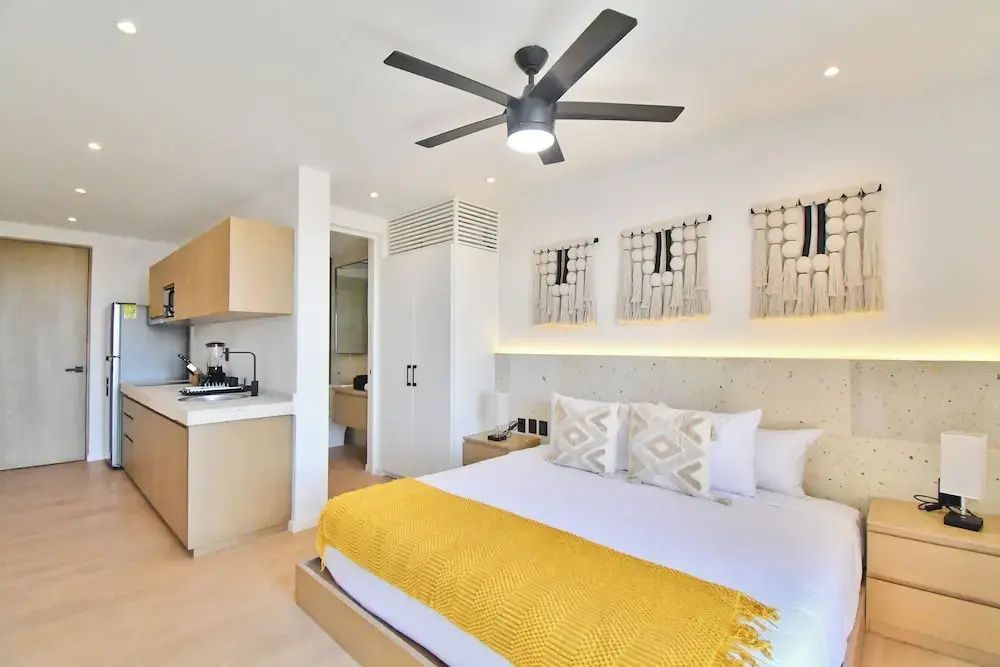Schlafzimmer mit Bett, Makramee-Wandbehängen, Deckenventilator und kleiner Küchenzeile.