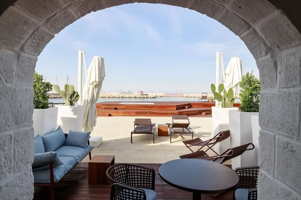 Terrasse mit Sitzmöbeln und Sonnenliegen unter einem Steinbogen mit Blick auf das Meer und den Hafen.