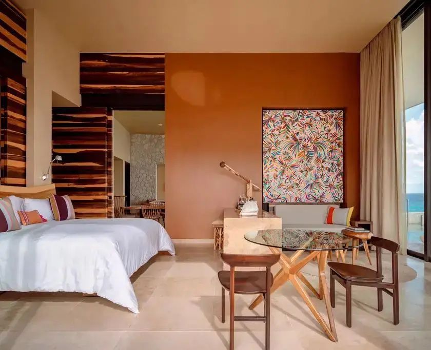 Hotelzimmer mit Doppelbett, hölzernem Esstisch, Schreibtisch und buntem Kunstwerk an der Wand.
