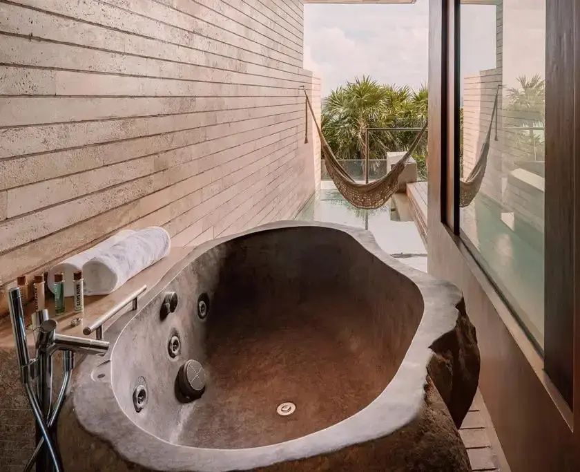 Freistehende Steinbadewanne auf der Terrasse mit Blick auf eine Hängematte und Grünpflanzen.