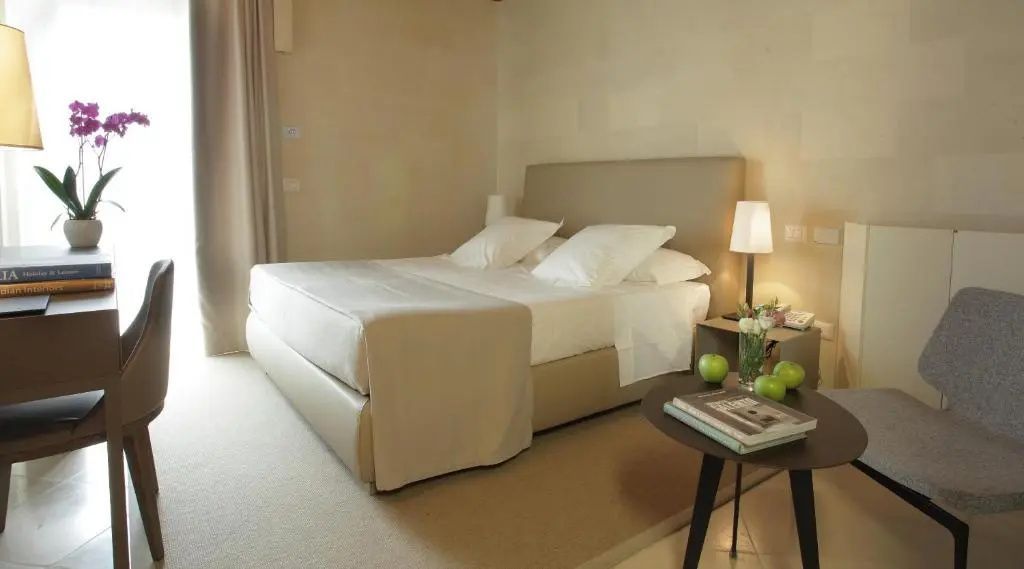 Doppelbett in hellem Hotelzimmer mit beigefarbenem Kopfteil, Nachttischlampe und Beistelltisch.