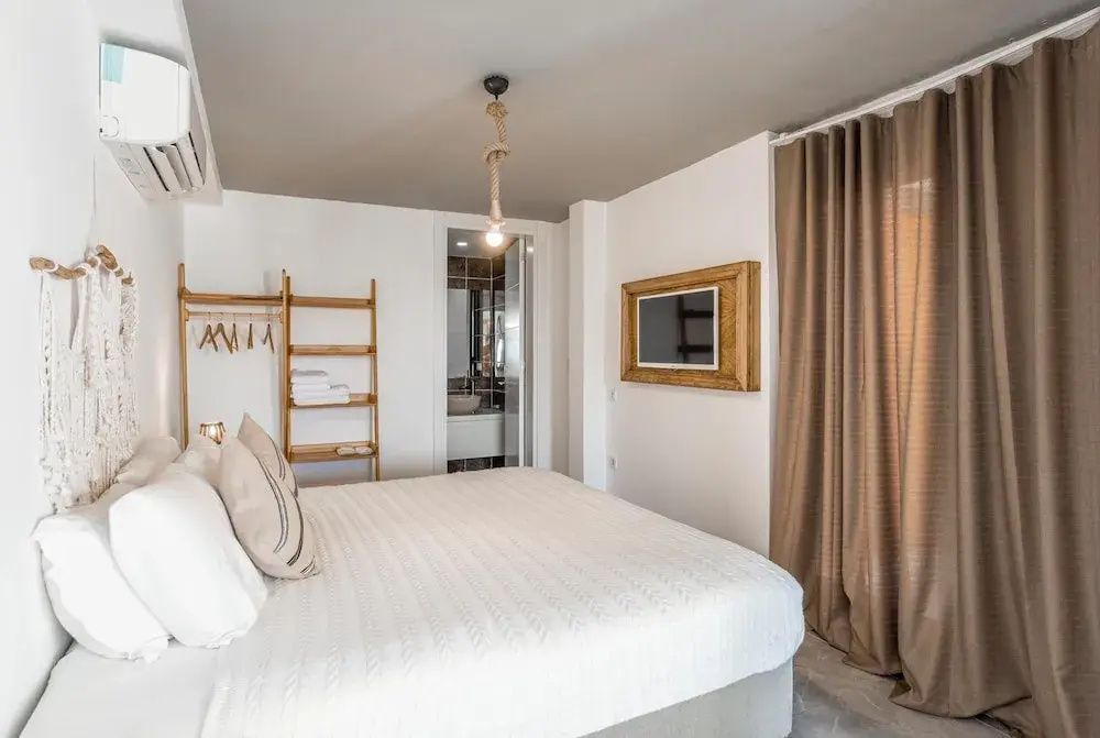Doppelbett mit weißer Bettwäsche, Holzregal, Fernseher an der Wand und Zugang zum Badezimmer.