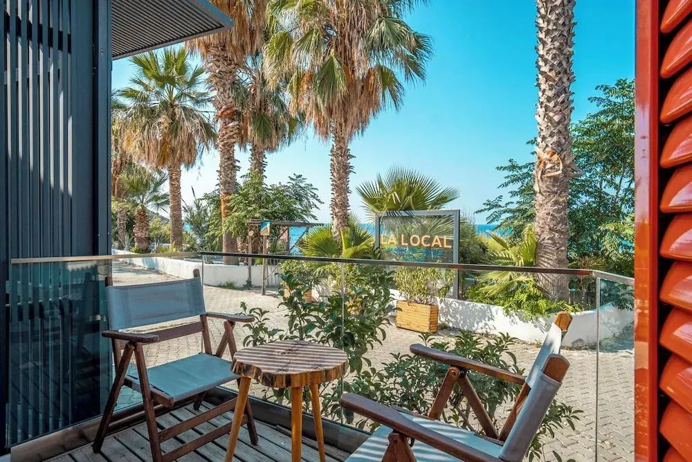 Holzterrasse mit zwei Stühlen und Beistelltisch mit Blick auf Palmen und das Meer.