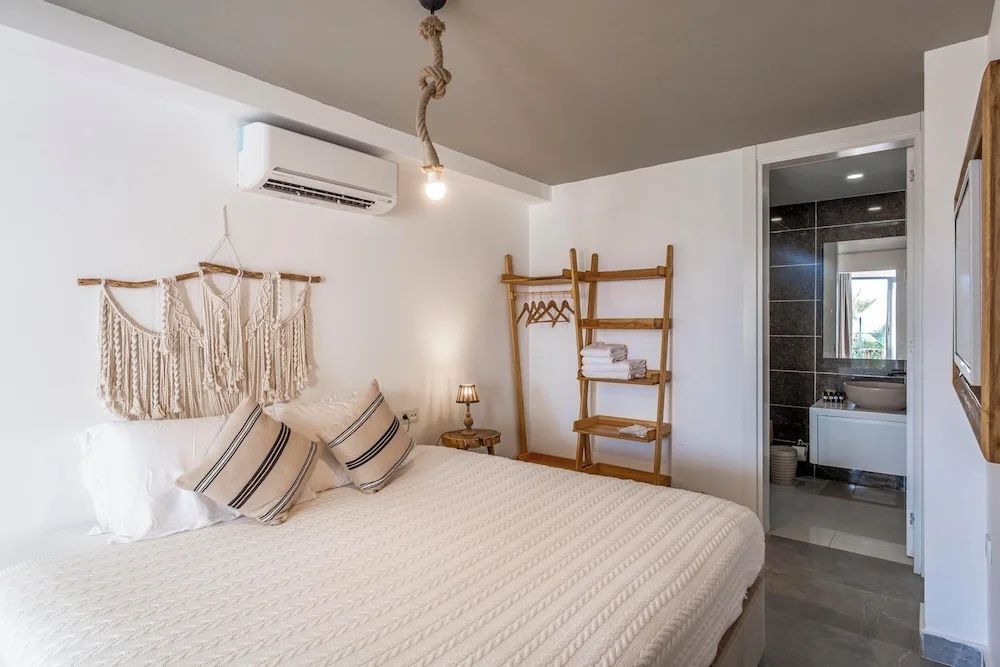Helles Hotelzimmer mit Doppelbett, Makramee-Wanddeko, Holzgarderobe und Blick ins Badezimmer.