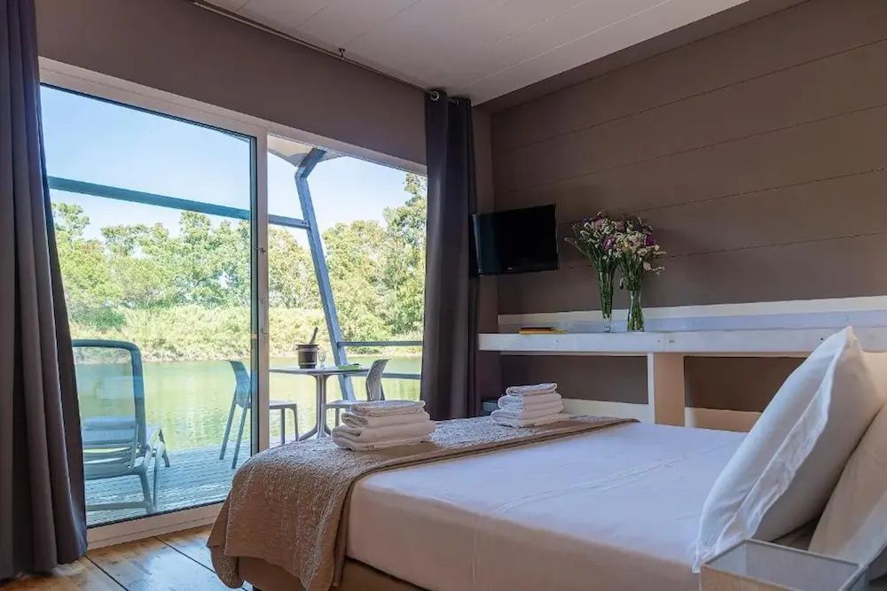 Helles Schlafzimmer mit Doppelbett, Fernseher und großer Glasfront zum See.