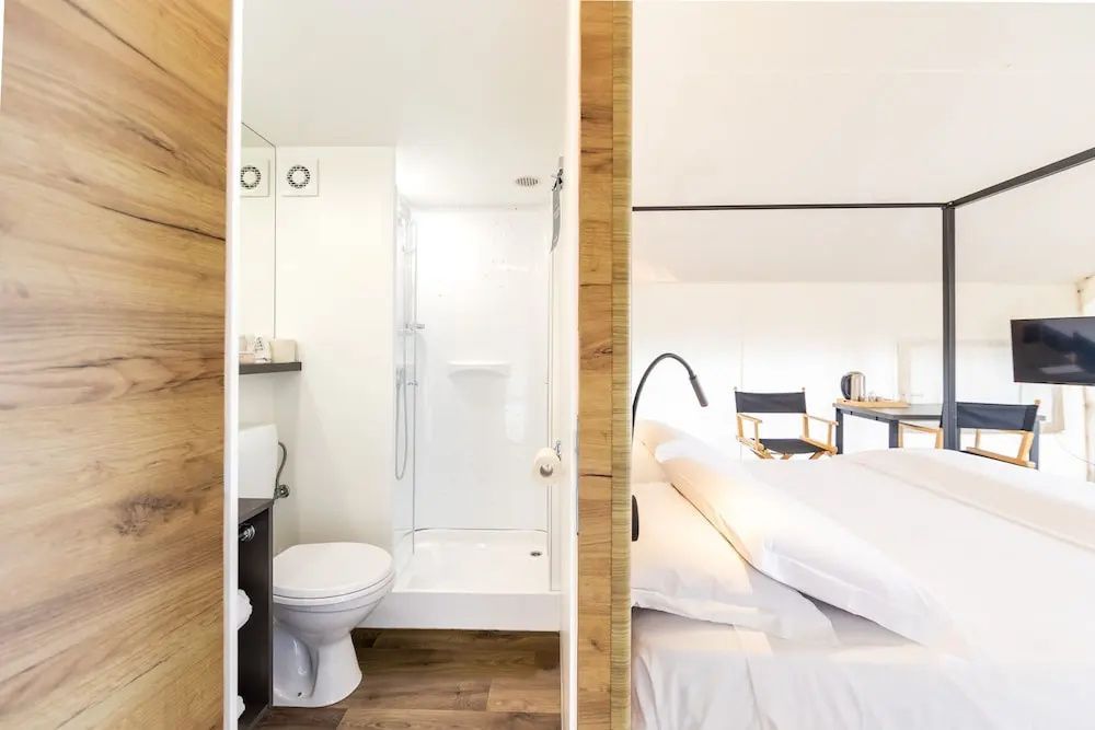 Blick auf ein Bett und das angrenzende, weiße Badezimmer mit Dusche und Toilette.