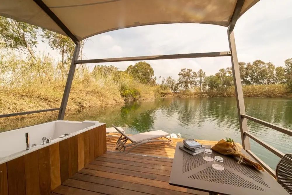 Blick von einer überdachten Holzterrasse mit Jacuzzi und Sonnenliege auf einen See.