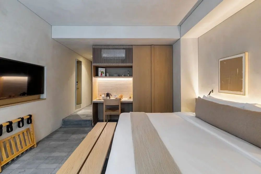 Hell gestaltetes Hotelzimmer mit Doppelbett, Holzbank, Einbauschrank und grau gefliestem Fußboden.