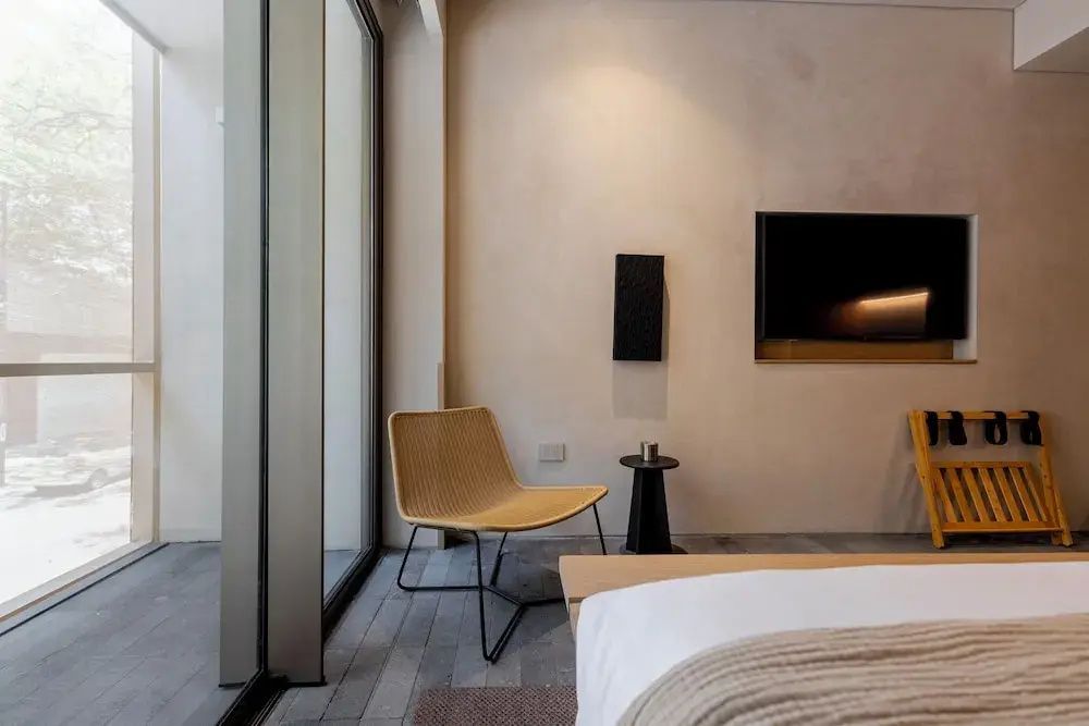 Sitzbereich im Hotelzimmer mit einem gewobenen Stuhl, beigefarbener Wand und einem flachen Fernseher.