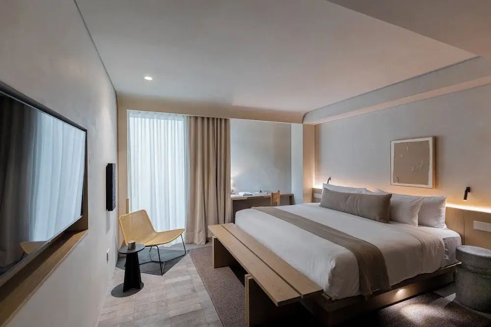 Übersichtliches Hotelzimmer mit Doppelbett, Sitzgelegenheit und hellem Vorhang.