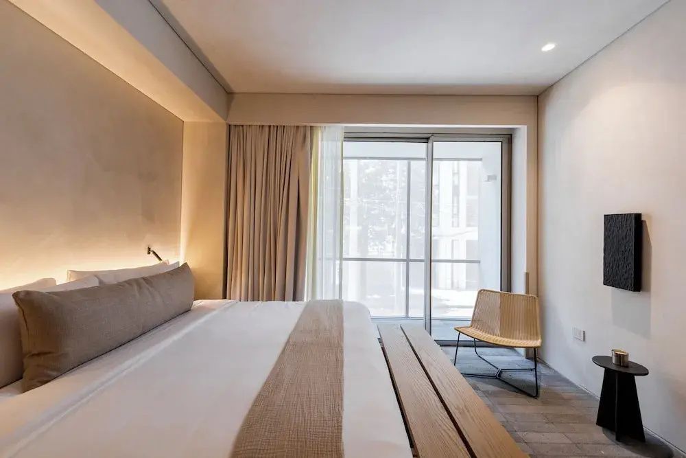 Helles Schlafzimmer mit Doppelbett, Holzbank und großer Glas-Schiebetür.