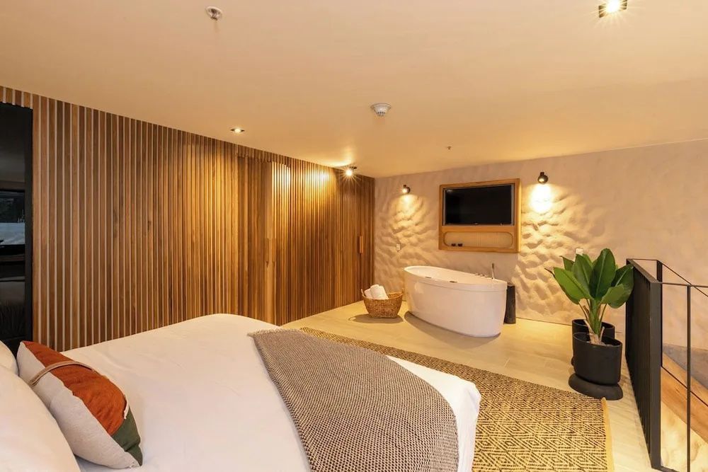 Schlafbereich mit Holzverkleidung, Bett, Fernseher an strukturierter Wand und Badewanne.