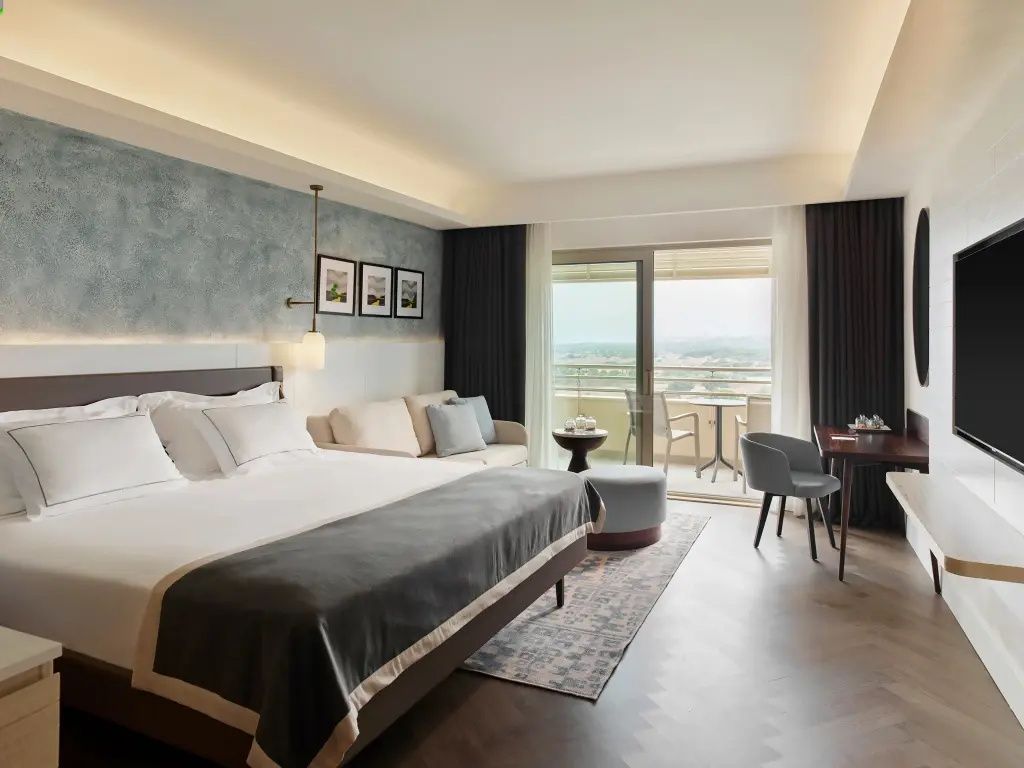 Helles Schlafzimmer mit Doppelbett, Couch, Schreibtisch und Fenstertür zum Balkon.