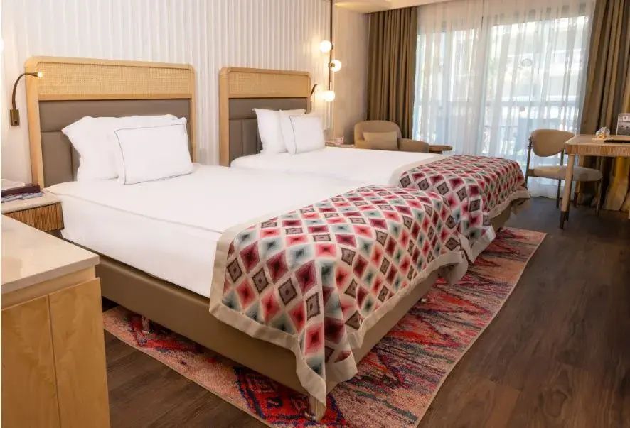 Helles Hotelzimmer mit zwei Einzelbetten, bunt gemusterten Läufern und Sessel am Fenster.