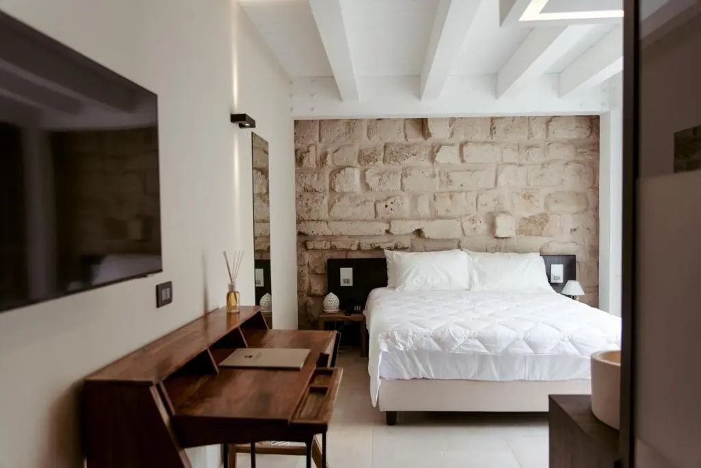 Schlafbereich mit Doppelbett vor einer Steinwand und Holzschreibtisch an der Wand.