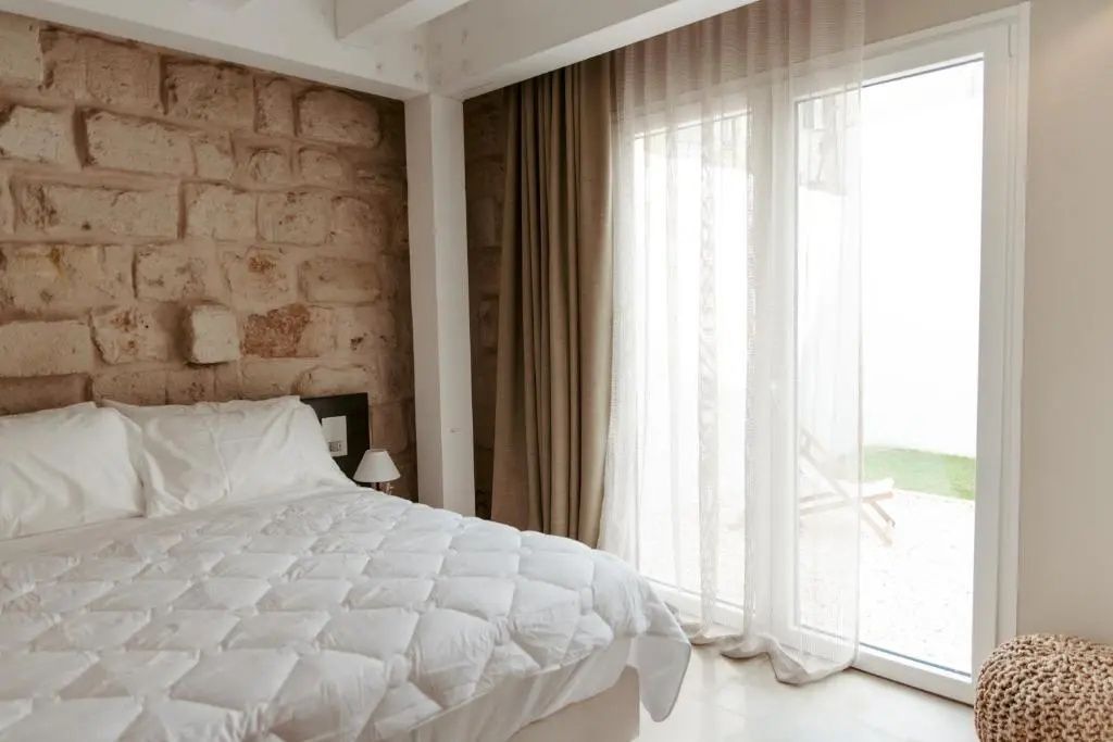 Hotelzimmer mit weißem Doppelbett, Steinwand und bodentiefen Fenstern zur Terrasse.