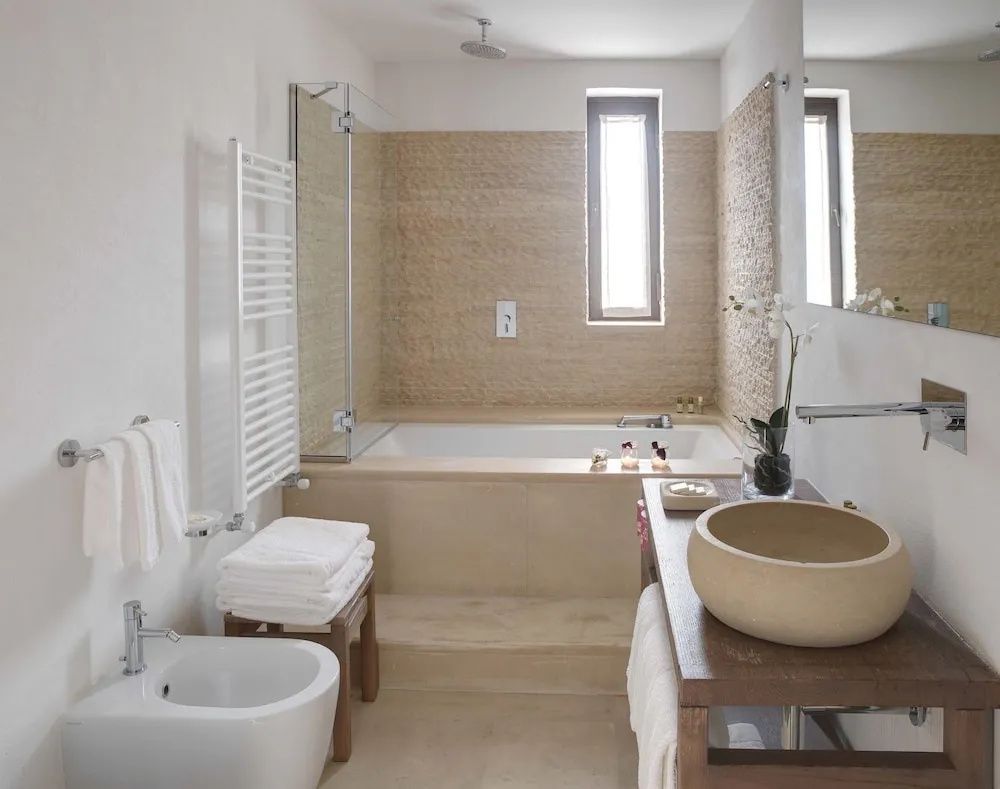 Badezimmer in Naturtönen mit tiefem Badewannenbereich, Wandfliesen aus Naturstein und Aufsatzwaschbecken.