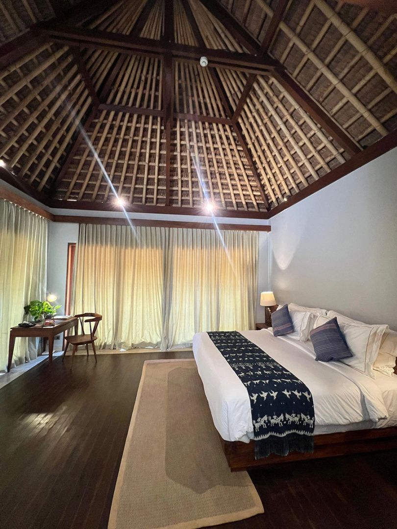 Schlafzimmer einer Villa mit großem Doppelbett, dunklem Holzboden und einer hohen Reetdachdecke.
