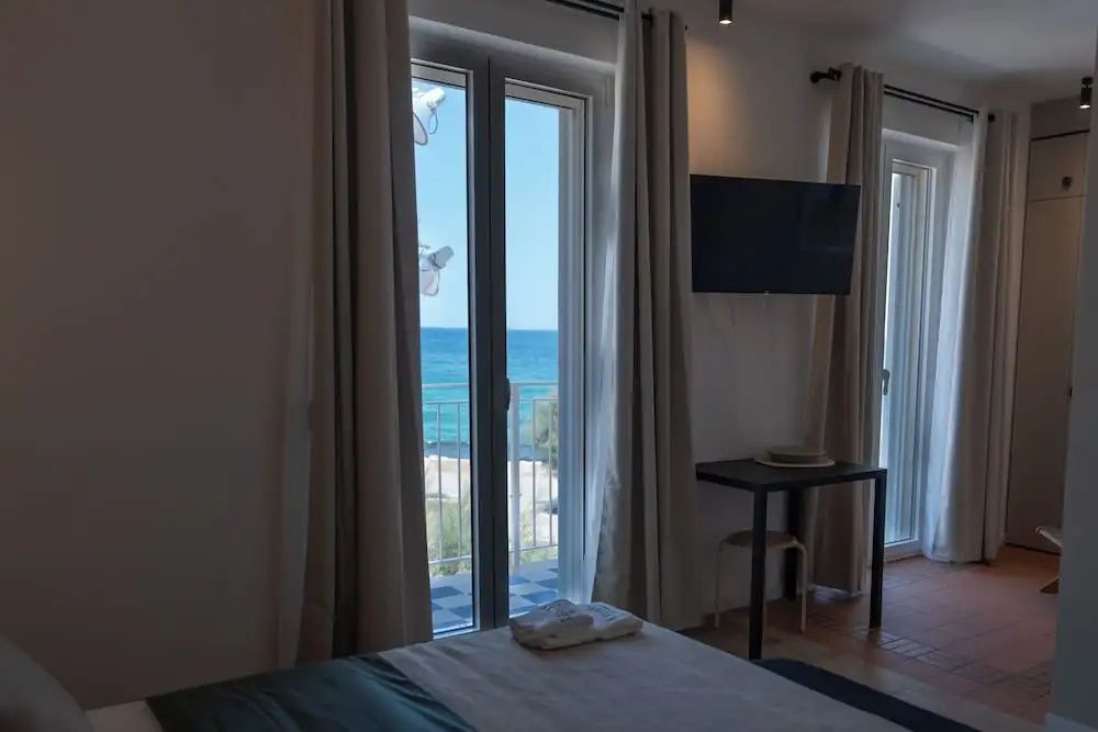 Blick aus dem Hotelzimmer durch eine Glastür auf das blaue Meer und den Strand.