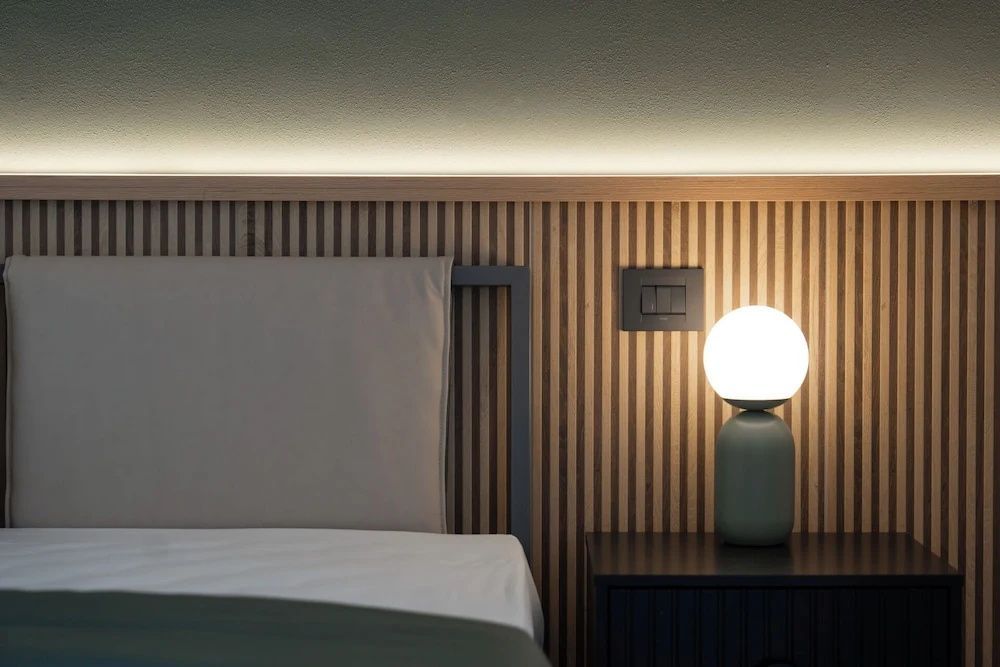 Kopfteil eines Bettes mit Holzlamellen, indirekter Beleuchtung und einer Kugel-Nachttischlampe.