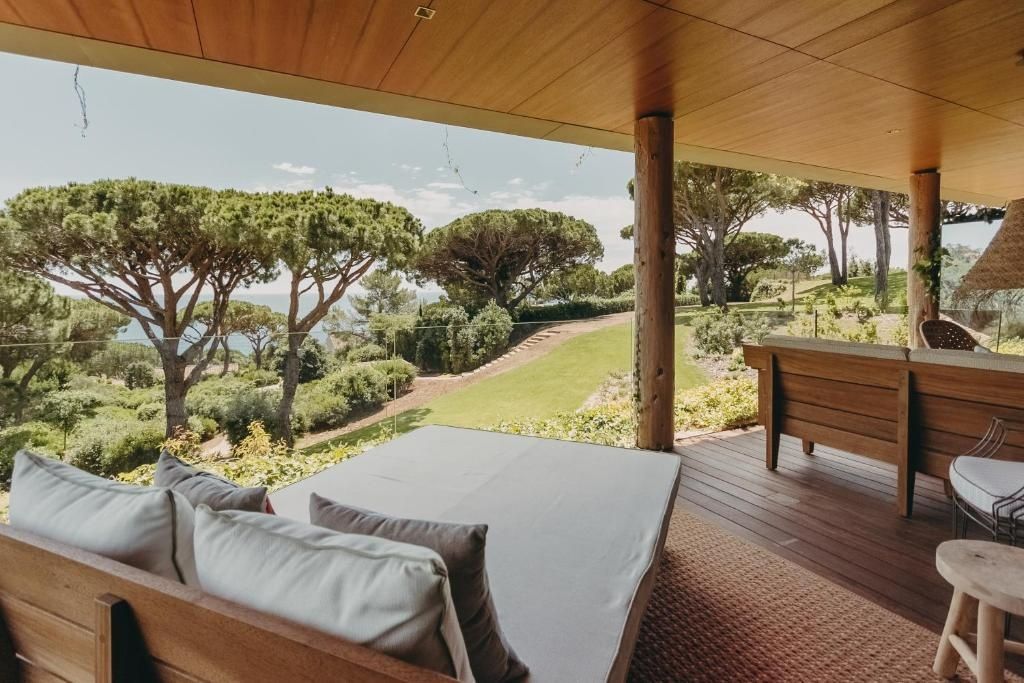 Überdachte Holzterrasse mit Tagesbett und Blick auf Pinienbäume und Meer.