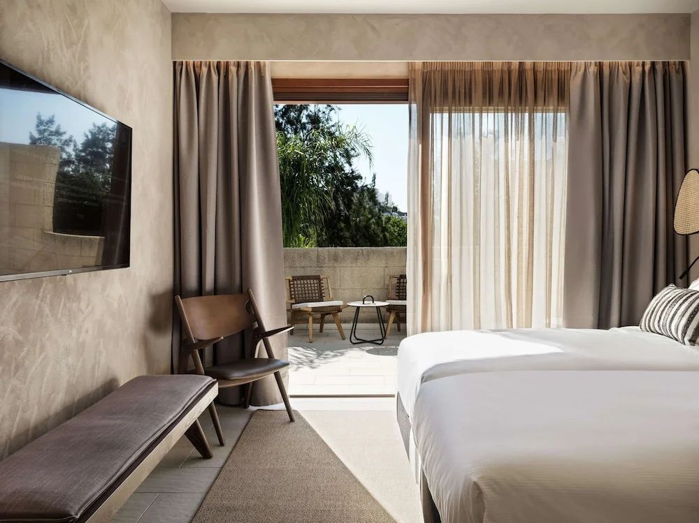 Hotelzimmer mit zwei Betten, Holzstuhl und geöffneter Balkontür zum Außenbereich mit Sitzgelegenheit.