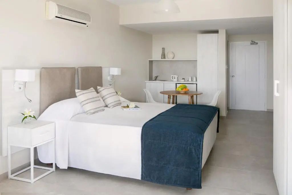 Helles Hotelzimmer mit Doppelbett, blauer Tagesdecke, Sitzbereich und kleiner Küchenzeile.