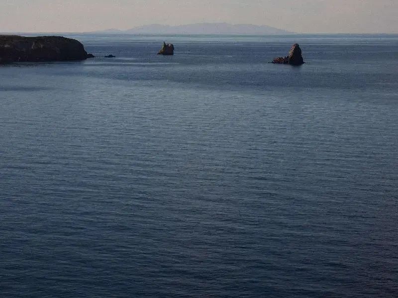 Weiter Blick über das ruhige blaue Meer auf Felsformationen und Inseln am Horizont.