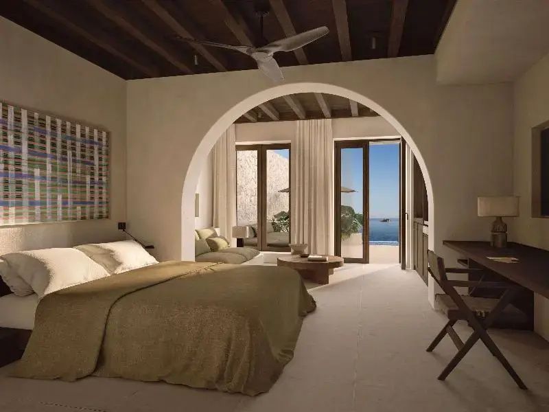 Doppelbett mit beiger Bettwäsche vor einem Rundbogen und Blick auf das Meer.