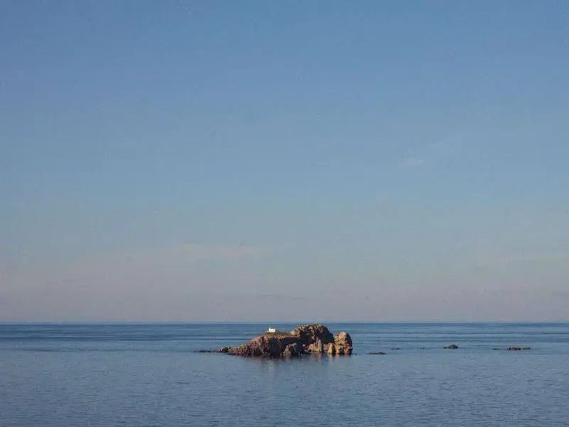 Blick über das blaue Meer auf eine kleine Felseninsel bei klarem Himmel.