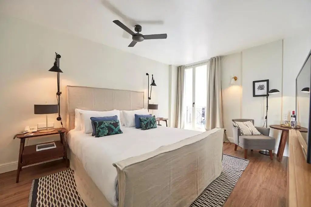 Doppelbett mit hellem Kopfteil, blauen Kissen, Deckenventilator und Sessel am Fenster.