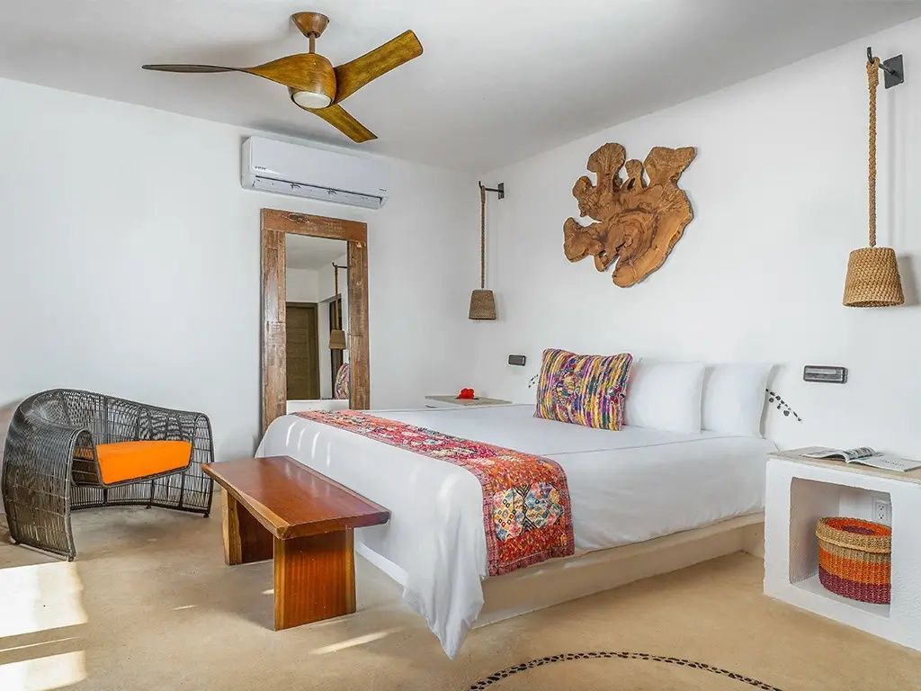 Heller Schlafraum mit Doppelbett, buntem Bettschal, Holzbank, Spiegel und Deckenventilator.