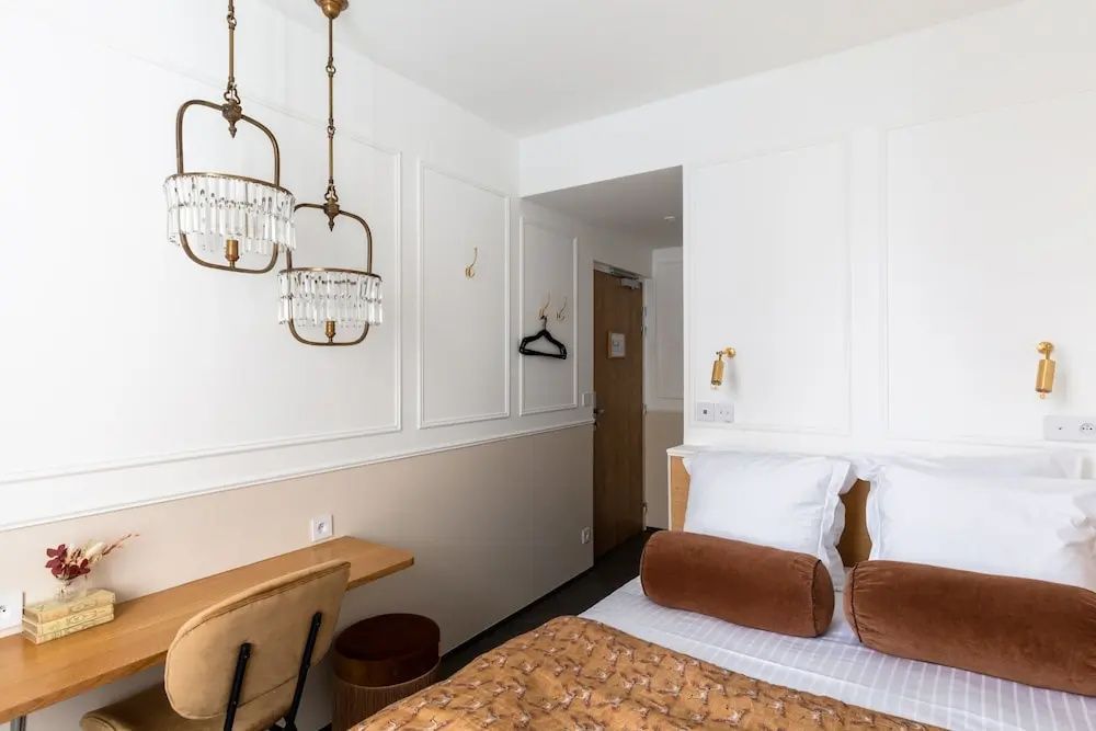 Helles Hotelzimmer mit Doppelbett, Holzschreibtisch und hängenden Kronleuchtern an weißer Wand.