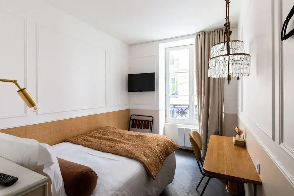 Blick in ein helles Zimmer mit Bett, Holzschreibtisch, Wandfernseher und hohem Fenster.