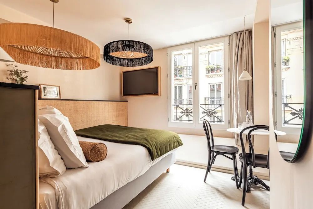 Blick in ein Zimmer mit Doppelbett, schwarzem Stuhl, Tisch und Fenster mit Blick auf die Stadt.