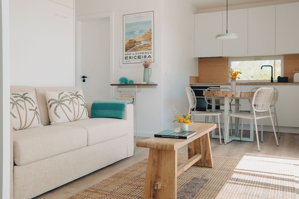 Heller Wohnbereich mit beigem Sofa, Holzbank, Holztisch und einer kleinen Küche im Hintergrund.