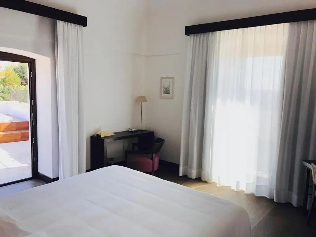 Schlafbereich mit weiß bezogenem Bett, dunklem Schreibtisch und Vorhängen am Fenster.